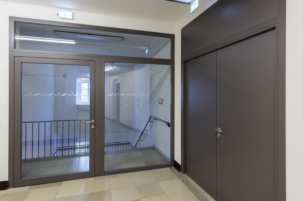 Durchsichtige Glastür mit modernem Rahmen, Zugang zu einem Raum mit Treppe und Geländer im Hintergrund.