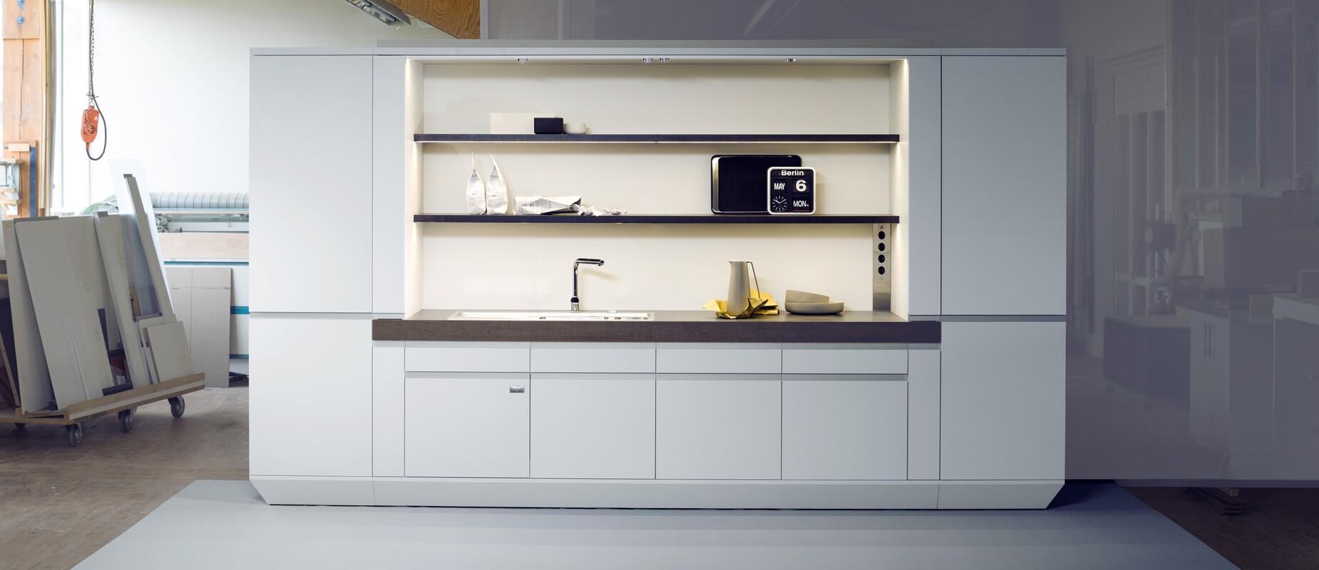 Moderne, weiße Küchenzeile mit Spüle, Regalen und Elektrogeräten in einem Werkstattumfeld.