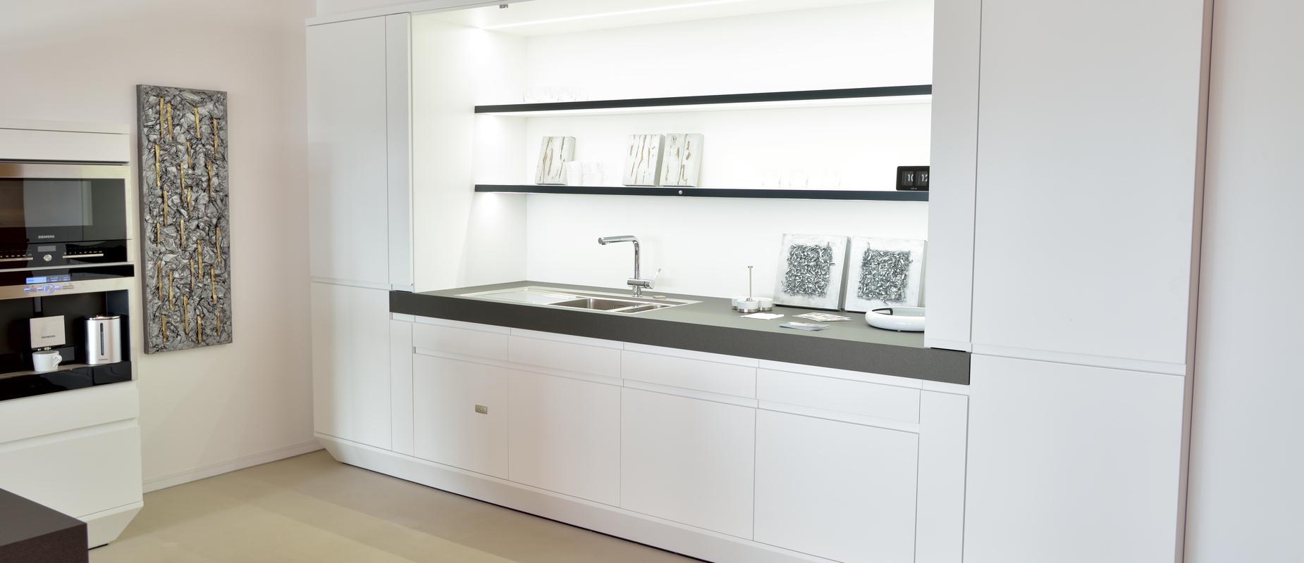 Moderne weiße Küche mit schwarzer Arbeitsfläche, Spüle und offenen Regalen, vor neutralem Hintergrund.