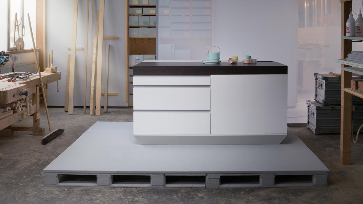 Moderne Kücheninsel mit Holzoberfläche, auf einem grauen Palettensockel in einem hellen Werkstattbereich.