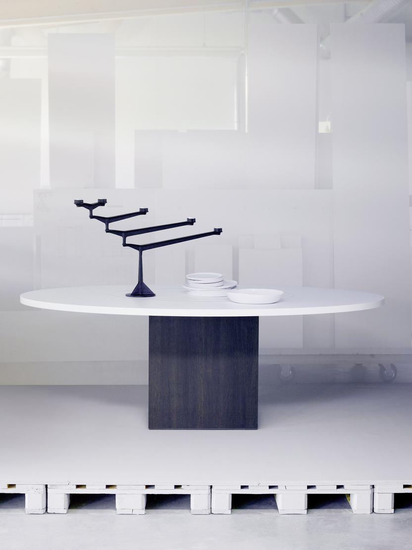 Kreisförmiger Tisch mit schwarzem Gestell und weißen Tellern, umgeben von minimalistischer, transparenter Raumgestaltung.