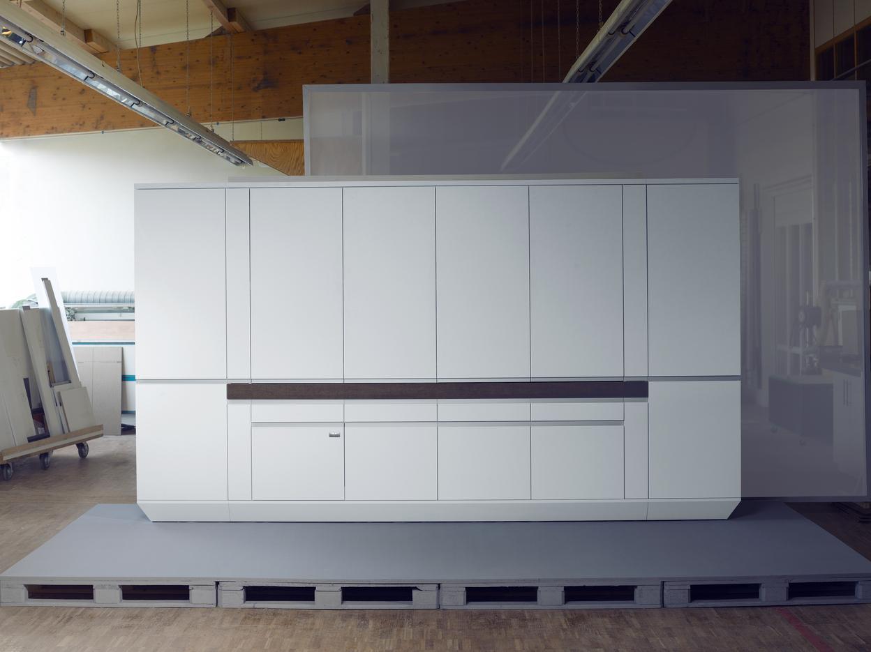 Moderne, weiße Küchenzeile auf einer grauen Plattform in einer hellen Werkstatt. Starke Linien und minimalistische Gestaltung.