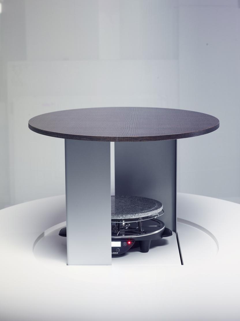 Runde Tischplatte aus Holz über einem grauen Heizgerät in einer modernen, minimalistischen Umgebung.