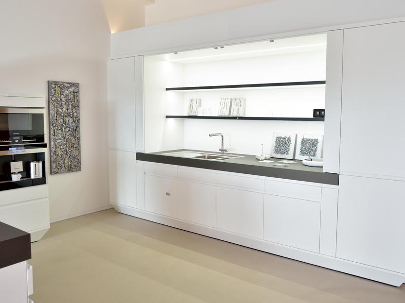 Moderne, minimalistische Küche mit weißen Schränken, schwarzer Arbeitsfläche und beleuchtetem Regal.