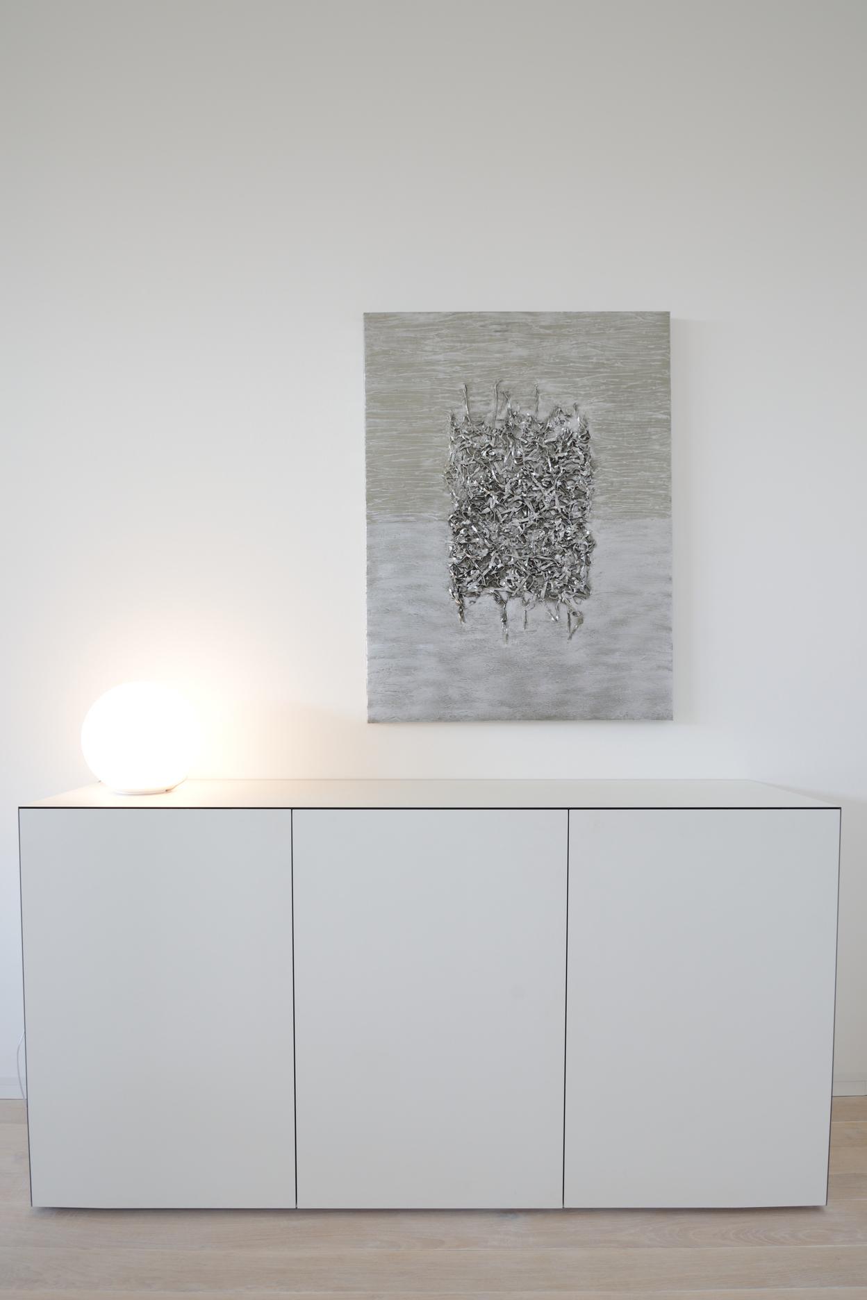 Weißer Sideboard mit drei Türen, beleuchtet von einer runden Lampe, im Hintergrund ein Wandbild in gedeckten Farben.