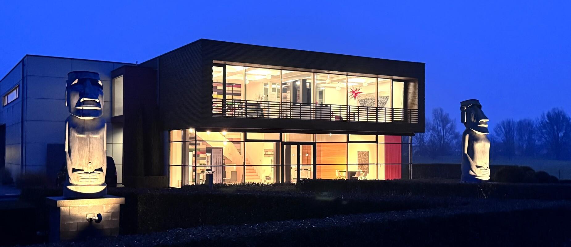 Moderne Architektur eines Gebäudes bei Dämmerung, flankiert von zwei Moai-Statuen in einem Garten.